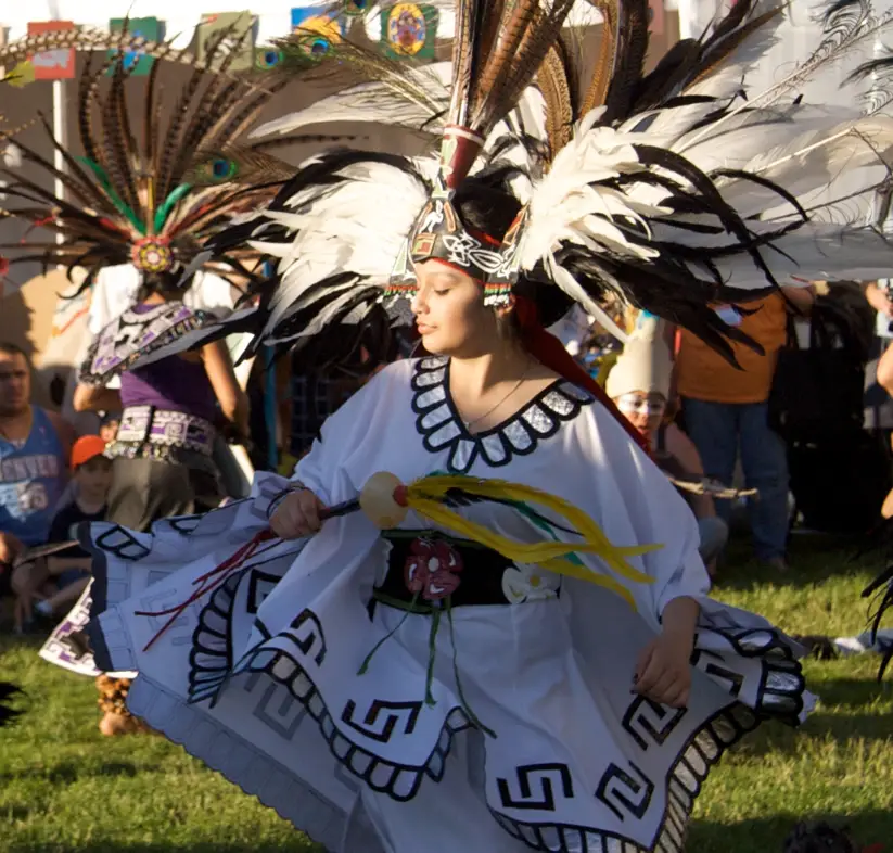 A dancer donning white garb and an ornate, feathered headdress twirls with eyes closed.